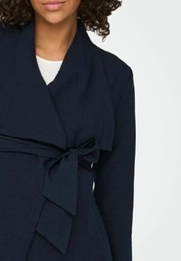 Donkerblauwe wikkelblouse met een strik in de taille, uitgevoerd in een gestructureerde stof en een diepe V-hals, die elegante, strakke lijnen toont.