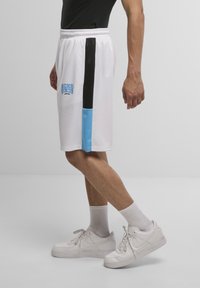 Pantaloni sportivi bianchi con dettagli neri e blu, caratterizzati da un logo sulla gamba sinistra. Indossati con sneakers bianche e calzini a coste.