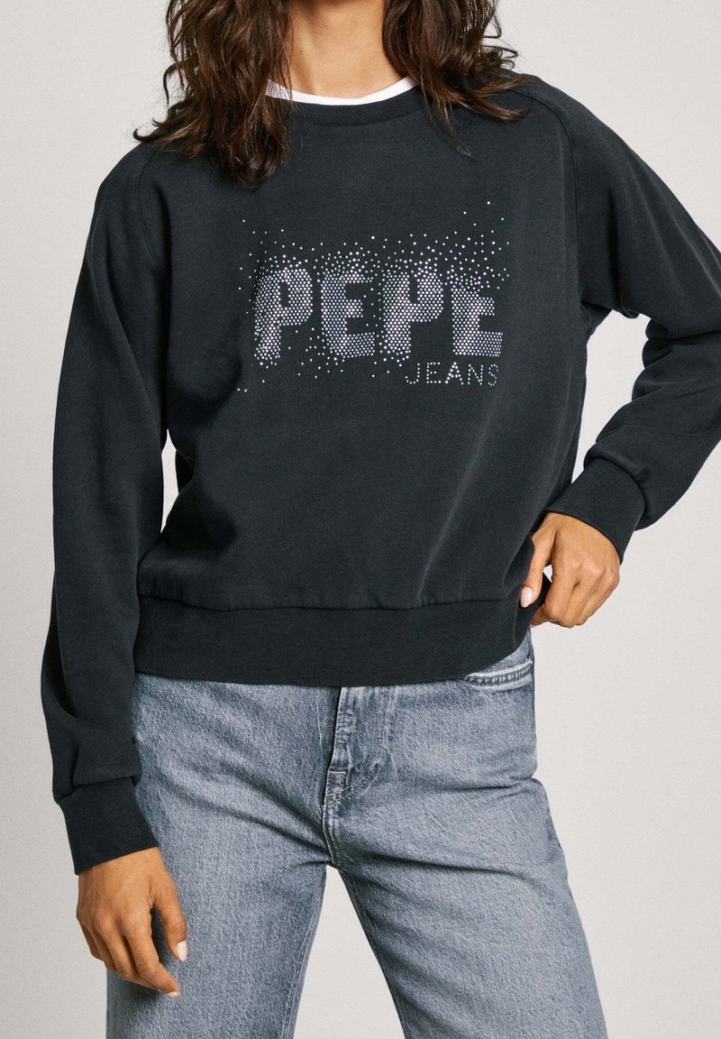 Femme portant un sweat-shirt noir à manches longues avec un texte blanc "PEPE JEANS" composé de petits points, associé à un jean bleu clair.