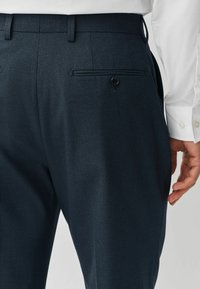 Navy trousers with a textured finish, featuring a back pocket secured by a button and a flat waistband. Paired with a white shirt.