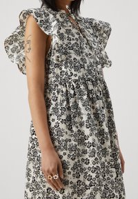 Květinové šaty v krémové a černé barvě, s volánovými rukávy, zapínáním na knoflíky a nařaseným pasem. Doplňky zahrnují květinové prsteny.