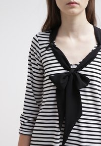 Svartvit randig topp med trekvartsärmar, med v-ringning och en stor svart rosett vid kragen. Mjuk tygkänsla.