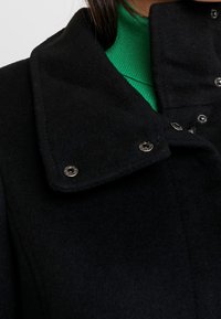 Černý vlněný kabát s velkým límcem, vyznačující se stříbrnými druky a texturovaným povrchem. Pod ním je vidět zelený pletený top.