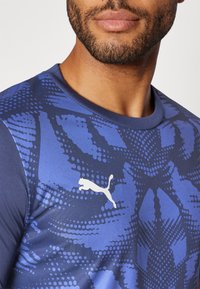 Kortermet blå sportskjorte med et teksturert design som har mørke og lyse blå mønstre. Hvit Puma-logo på brystet.