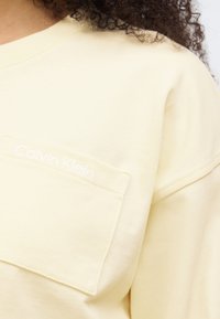 Ανοιχτό κίτρινο πουλόβερ με τσέπη που διαθέτει λευκή κεντημένη επιγραφή "Calvin Klein". Μαλακή υφή, άνετη εφαρμογή και εμφανείς ραφές.