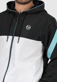 Capuchonjack met een zwart bovenstuk en een wit onderstuk, met teal accenten op de capuchon en de mouwen. Ritsclosure en logo-detail.