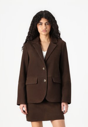 Veste en cuir brun suede avec un col cranté, deux poches avant et un bouton unique, associée à une jupe assortie.