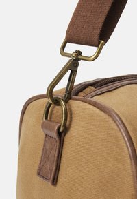 Un sac en tissu brun avec un attachement en clips en laiton, des accents en cuir et des détails cousus. Le design comprend une fermeture éclair et une sangle plate.