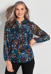 Donna matura con capelli grigi che sorride, indossando orecchini a cerchio dorati, una blusa a fantasia multicolore, pantaloni neri e un bracciale dorato.