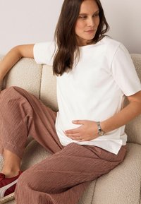 Weißes Baumwoll-T-Shirt, locker sitzende rot-weiß karierte Hose, graue Uhr, sitzt auf einem strukturierten beigen Sofa, die Hand auf dem Bauch ruhend.