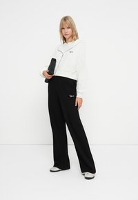 Bela zip-up kapuca z ribasto teksturo in črnima logotipom v kombinaciji s širšimi črnimi hlačami in obutvijo s srebrnimi detajli.