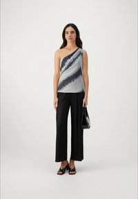 Donna che indossa un top grigio e nero a spalla unica, pantaloni larghi neri, tacchi aperti neri, mentre tiene una borsa nera.
