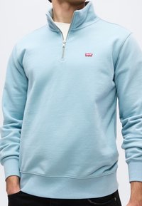 Homme portant un sweat-shirt bleu clair à fermeture éclair quart de zip avec logo Levi's rouge sur la poitrine gauche, un sous-t-shirt blanc et un pantalon noir.