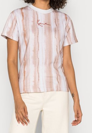 Kobieta ubrana w biało-jasnobrązowy pasiasty t-shirt z krótkim rękawem z wyhaftowanym na czerwono napisem „Kani” na piersi, zestawiony z białymi spodniami.