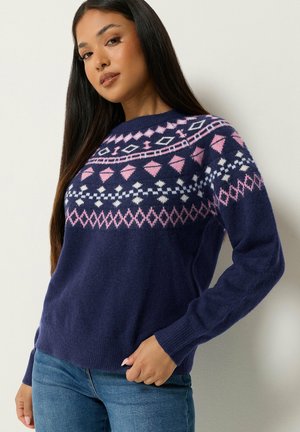 Mujer que lleva un suéter azul marino con patrones geométricos rosas y blancos en el pecho y los brazos superiores, combinados con vaqueros azules.