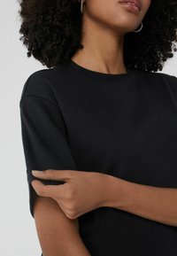 Czarna krótka koszulka z okrągłym dekoltem, o luźnym kroju i miękkiej fakturze. Ręka dotyka rękawa w celu regulacji.