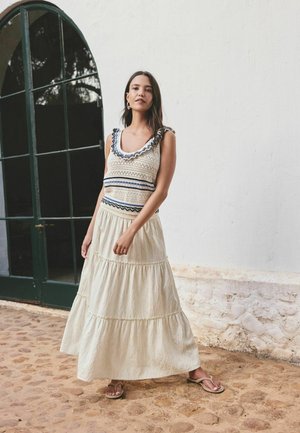 Femeie stând pe podea de piatră cubică, purtând o rochie crem fără mâneci, cu guler volanat și fustă în straturi, lângă o fereastră mare în arcadă.