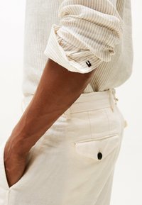Camisa a rayas beige con mangas enrolladas, que presenta una pequeña etiqueta con logo. Pantalones de corte relajado en un color claro, con un bolsillo trasero y un detalle de botón.