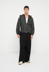 Siv zip-up jopič z kapuco, elastičnimi manšetami in dvema sprednjima žepoma; nošen preko bele majice in v kombinaciji s črnimi kvadratnimi širokimi hlačami.