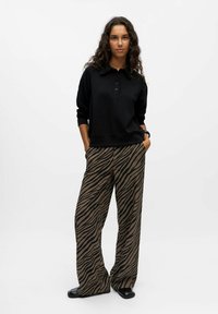 Schwarzes Poloshirt mit Knöpfen, kombiniert mit hochtaillierten braunen Hosen, die ein auffälliges schwarzes Zebra-Streifenmuster aufweisen. Strukturiertes Gewebe.
