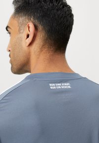 Camiseta atlética gris de mangas cortas, con texto blanco en la parte posterior que dice "NUR EINE STADT. NUR EIN VEREIN." Textura suave.