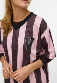 Trumparankovė sportinė marškinėliai su vertikaliomis tamsiai rudos ir šviesiai rožinės spalvos juostelėmis, juodu V formos logotipu ant krūtinės ir juodu V formos kaklu.