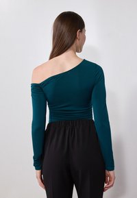 Haut teal à manches longues avec un design drapé, présentant un col asymétrique et des détails froncés. Assorti à un pantalon noir à taille élastique.