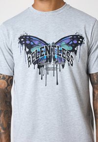 Camiseta de algodón gris con un gran gráfico de mariposa multicolor con gotas negras, con el texto "RELENTLESS" y "Closure London."