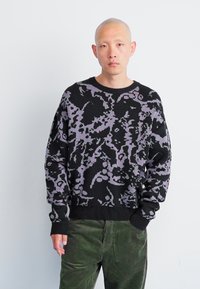 Pulover negru cu un model abstract purpuriu, gât rotund, manșete și tiv cu nervuri. Asortat cu pantaloni din corduroy verzi.