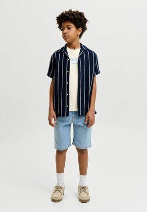 Niño con cabello rizado que lleva una camisa azul marino de manga corta con botones, camiseta gráfica color crema, pantalones cortos de mezclilla claros, calcetines blancos y zapatos beige, de pie sobre fondo blanco.