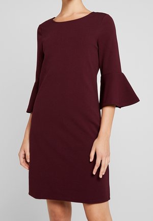 Kobieta w ciemnoczerwonej sukience do kolan z rozkloszowanymi rękawami o długości 3/4 i okrągłym dekoltem, stojąca na tle jasnoszarej, jednolitej ściany.
