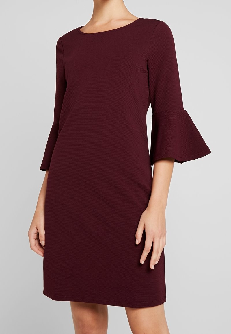 Femme portant une robe bordeaux foncé longueur genou avec des manches cloches trois-quarts et un col rond, debout devant un fond uni gris clair.