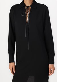 Robe noire à manches longues avec un col, présentant un décolleté en forme de clé et des liens en ruban satiné, fabriquée dans un tissu lisse avec une texture minimale.