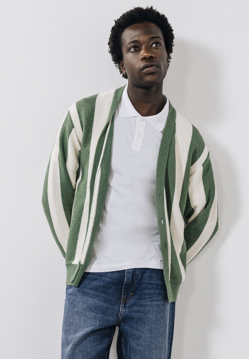Grüner und cremefarbener gestreifter Cardigan mit einer weichen Textur, Knopfverschluss und lockerer Passform, getragen über einem weißen Polohemd und blauen Jeans.