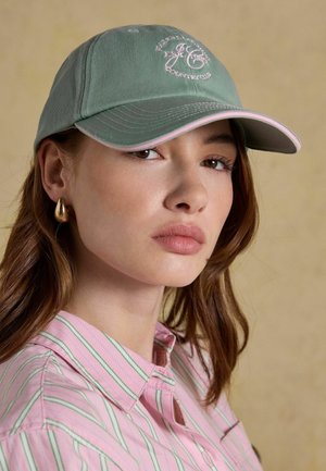 Mladá žena so svetlo hnedými vlasmi má na sebe zelenú šiltovku s ružovým lemovaním, zlaté kruhové náušnice a ružovo pruhovanú košeľu na gombíky na béžovom pozadí.