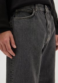 Jeans en denim gris foncé avec une texture légèrement délavée, un design à cinq poches et des accents de couture jaunes le long des coutures.