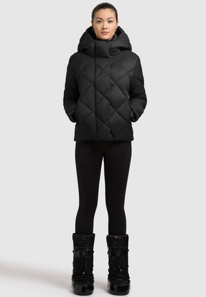 Giacca a vento nera con motivo a diamante, colletto alto e cappuccio. Indossata con leggings neri e stivaletti da neve neri e lucidi.