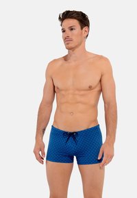 Shorts de bain bleus avec un motif en diamants, fermeture à cordon de serrage et design ajusté. Texture lisse et longueur mi-thigh.