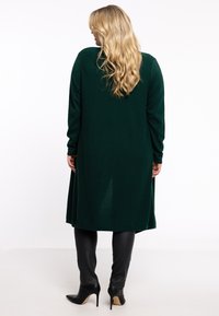Long gilet en maille verte avec des manches longues, présentant un design fluide, associé à un pantalon noir ajusté et des chaussures à talons pointus noires.