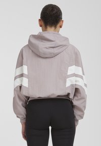 Personen står med ryggen til iført en cropped mauve hættetrøje med hvide striber på ærmerne og sorte tætsiddende leggings mod en ensfarvet baggrund.