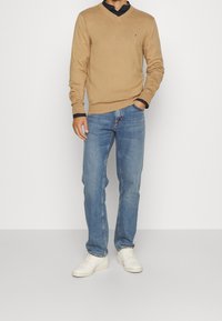 Beżowy sweter z dekoltem w serek, teksturowany materiał, czarna koszula pod spodem, niebieskie dżinsy i białe sneakersy. Posiada małe logo na swetrze.