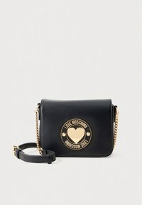Svart skulderveske i kunstskinn med en rund gullemblem som viser et hjerte og merkenavn. Inkluderer en gullkjede stropp og klafflukking.