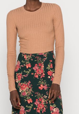 Lichtbruine geribbelde langemouwen crop top gecombineerd met een groene bloemenrok met rode en gele bloemen, waardoor een contrasterende outfit ontstaat.