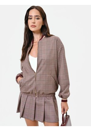 Donna che indossa una giacca marrone a quadri con zip e gonna plissettata abbinata, tenendo una piccola borsa bordeaux, con lunghi capelli scuri.