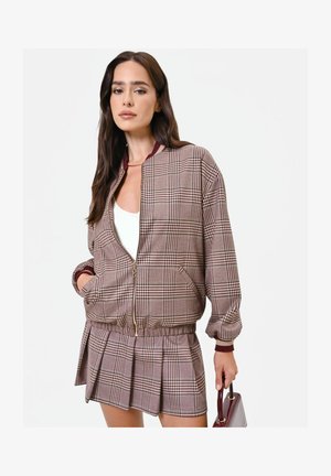 Donna che indossa una giacca marrone a quadri con zip e gonna plissettata abbinata, tenendo una piccola borsa bordeaux, con lunghi capelli scuri.