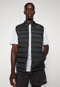 Gilet rembourré noir avec un col haut, fermeture éclair à l'avant et poches latérales ; logo sur la poitrine gauche ; associé à un t-shirt blanc.