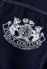 Λευκό κεντητό εμβλημα Juicy Couture με δύο σκύλους, ένα στέμμα και το κείμενο "Born in the Glamorous USA" σε σκούρο τζιν ύφασμα.