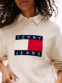 Beige sweater with ribbed neck, featuring a rectangular logo in navy, white, and red at the center. Soft texture, relaxed fit.