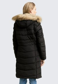 Lang zwarte pufferjack met een geborduurd ontwerp en een nepbont capuchon. Heeft een getailleerde pasvorm en reikt tot halverwege de dijen.