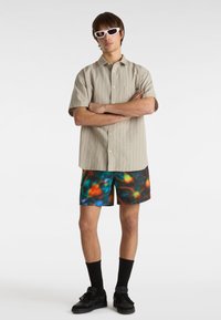 Kurzärmliges, gestreiftes beiges Hemd, farbenfrohe gemusterte Badeshorts, schwarze Socken, schwarze Sneaker und moderne weiße Sonnenbrille.
